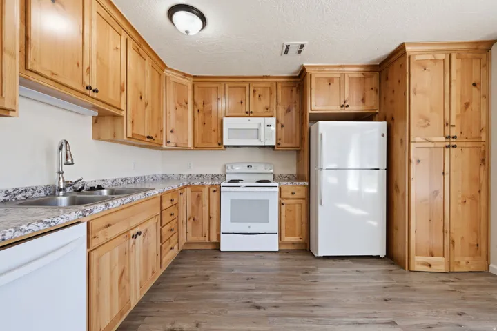 Kitchen featuring wood-finish cabinetry, light-toned countertops, a double basin stainless steel sink, wood-finish flooring, and white appliances