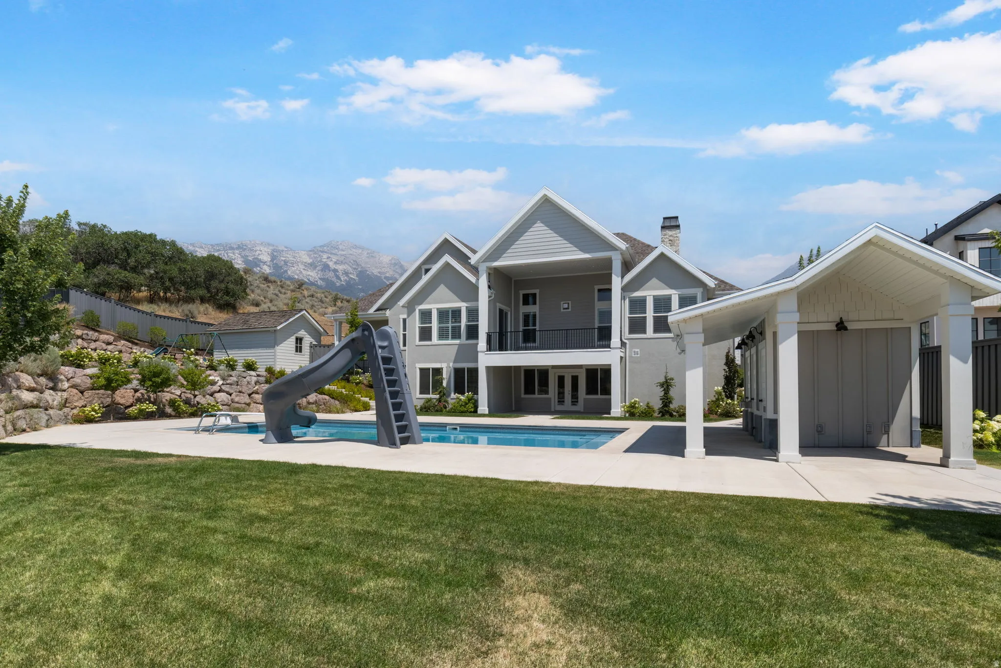 Expansive backyard featuring a manicured lawn, a swimming pool with a slide, a covered outdoor structure, and a retaining wall with landscaping