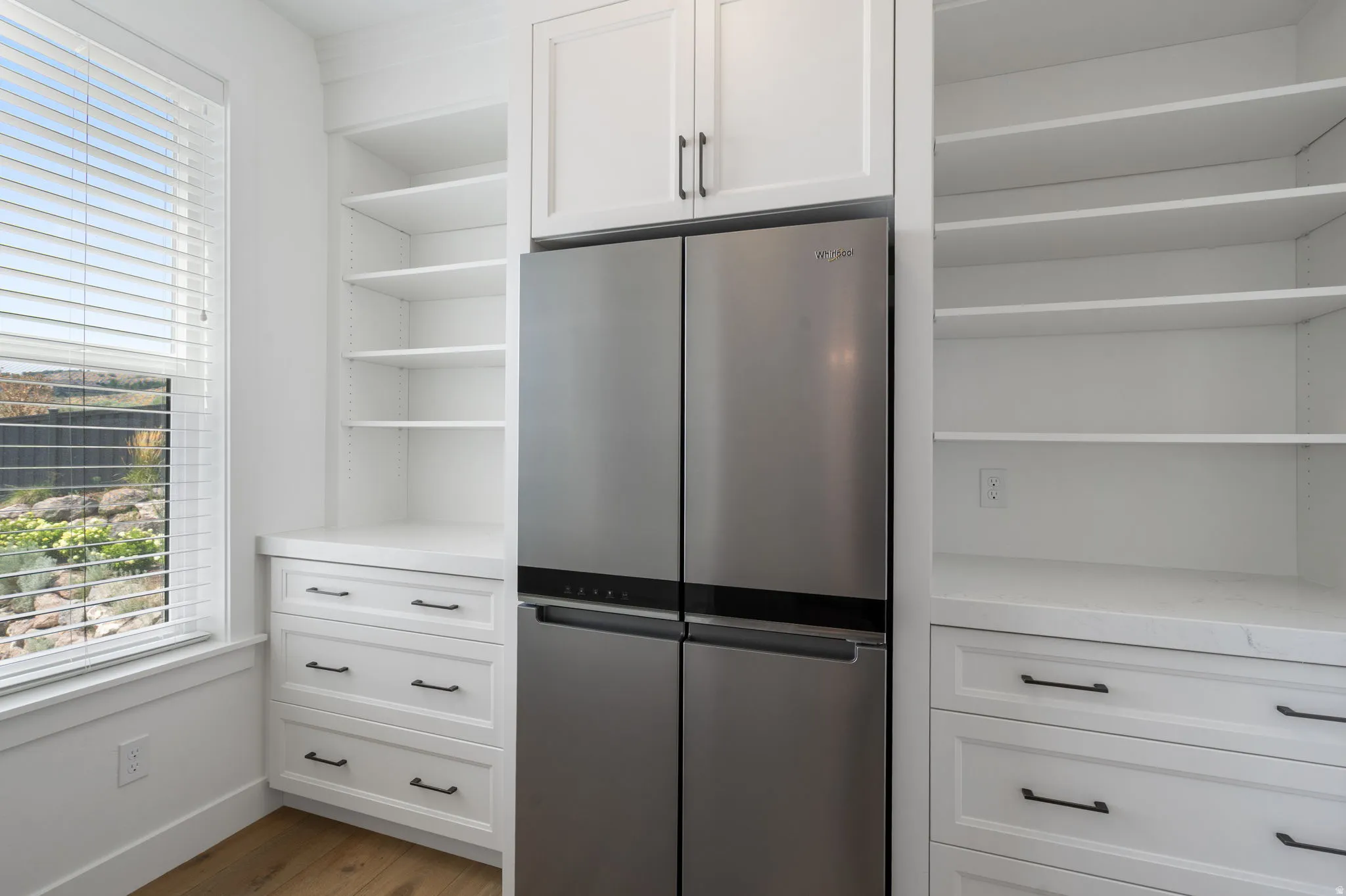 Stainless steel French door refrigerator, white built-in cabinetry, white shelving, white drawers with dark hardware, and wood-finish flooring