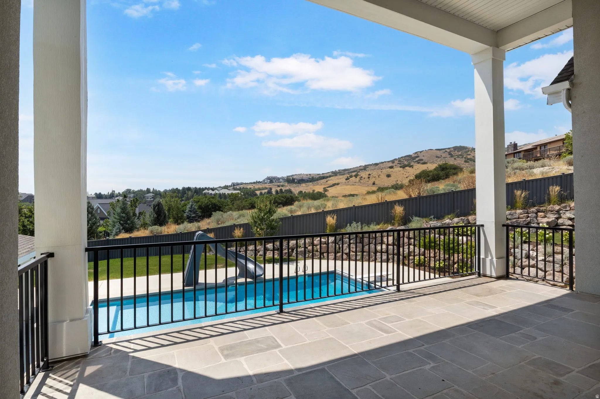 Covered patio with stone tile flooring, black metal railing, and an outdoor swimming pool featuring a slide