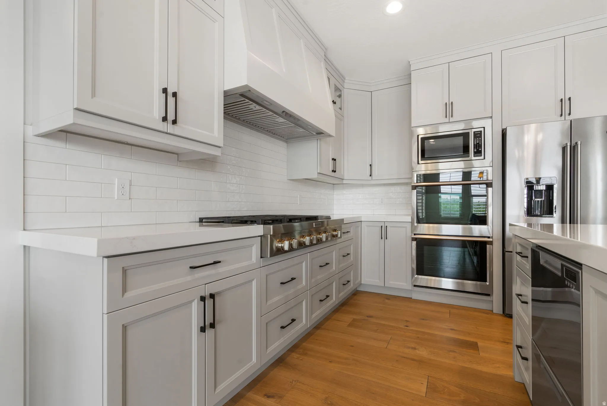 White shaker cabinetry with black hardware, white subway tile backsplash, light-toned countertops, professional-grade gas range, and wood-finish flooring