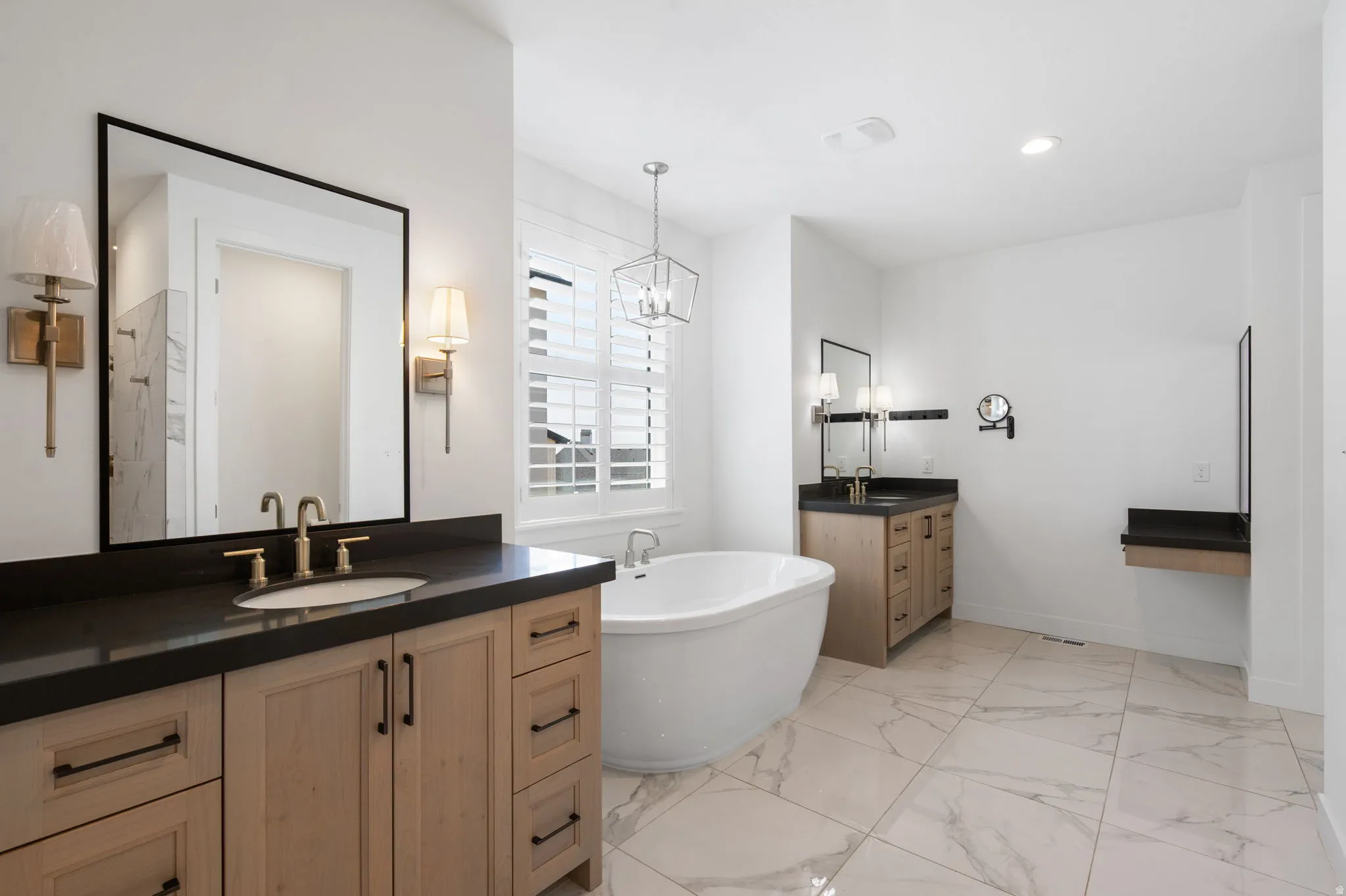 Spacious bathroom featuring a freestanding tub, dual vanities with dark countertops, wood-finish cabinetry, and large format marble-look tile flooring