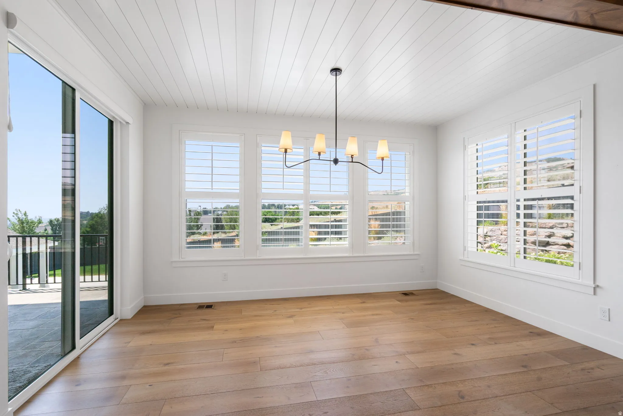 Wood-finish flooring, white shiplap ceiling, contemporary chandelier, plantation shutters, and sliding glass door