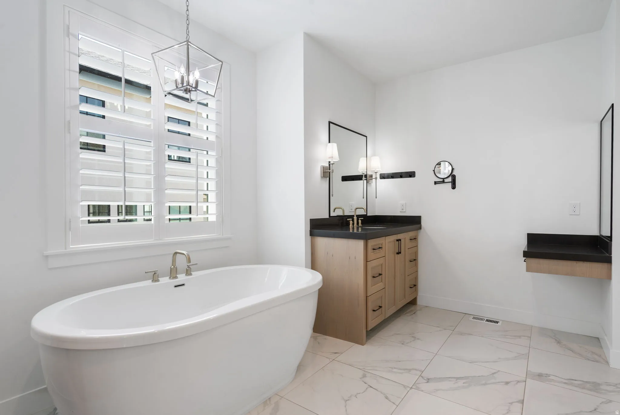 Freestanding bathtub with brushed nickel fixtures, plantation shutters, marble-look tile flooring, light wood-finish vanity with dark countertop, and decorative pendant lighting