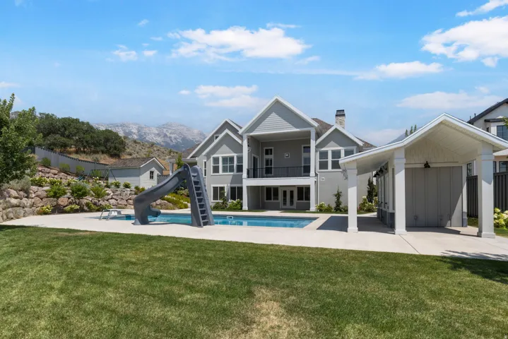 Expansive backyard featuring a manicured lawn, a swimming pool with a slide, a covered outdoor structure, and a retaining wall with landscaping