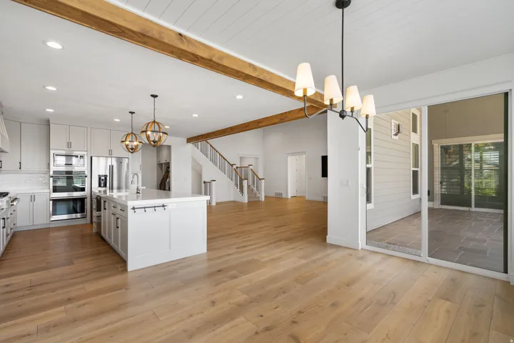 Open-concept living space with wood-finish flooring, exposed ceiling beams, a white kitchen island with seating, a full suite of stainless steel appliances, and a sliding glass door leading to an outdoor patio