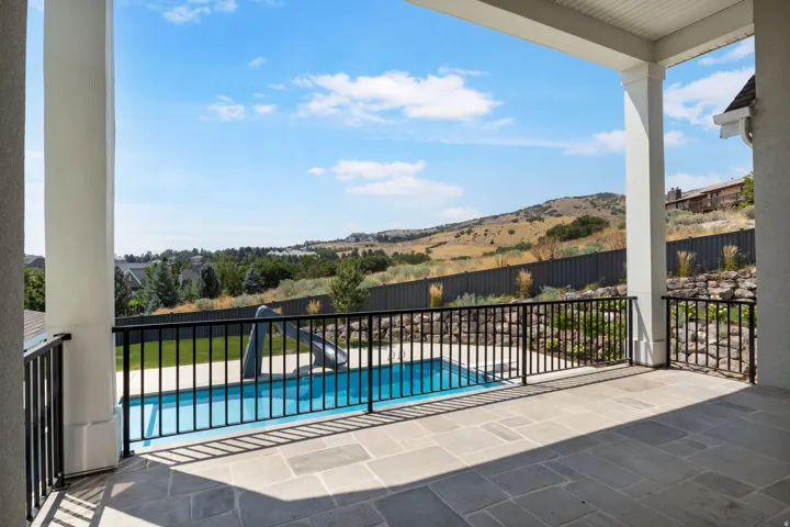 Covered patio with stone tile flooring, black metal railing, and an outdoor swimming pool featuring a slide