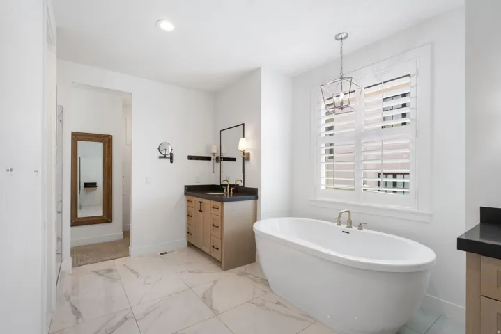 Freestanding soaking tub, plantation shutters, white tile flooring, wood-finish vanity with dark countertop, and brushed nickel fixtures