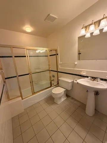 Pedestal sink with brass-finish fixtures, a full bathtub with a sliding glass enclosure, white tile flooring, and a vanity light fixture