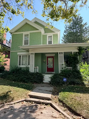 Gabled roofline, ornamental shingle siding, covered front porch with white columns, red front door with an upper window panel, and foundation plantings