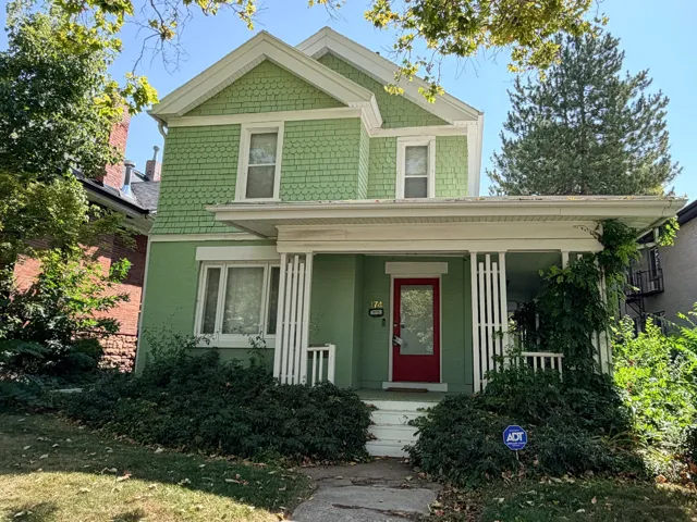 Gabled roofline, green shake siding, contrasting white trim, covered front porch with vertical wood railings, and a red entry door