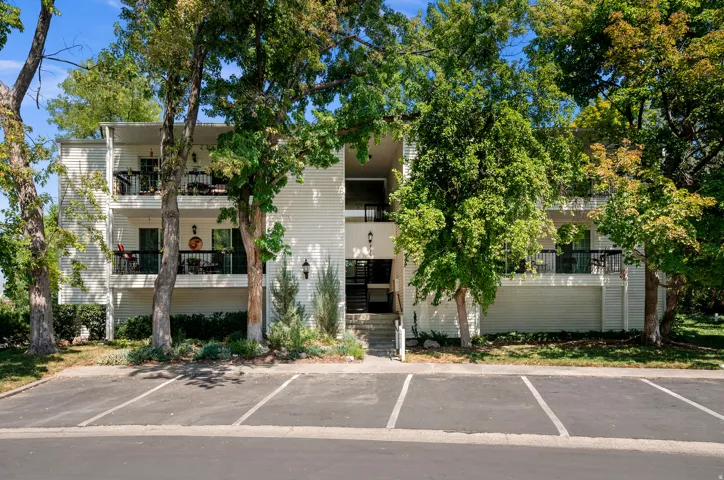 Two-story building exterior with vertical siding, multiple balconies, a central entryway, and mature trees