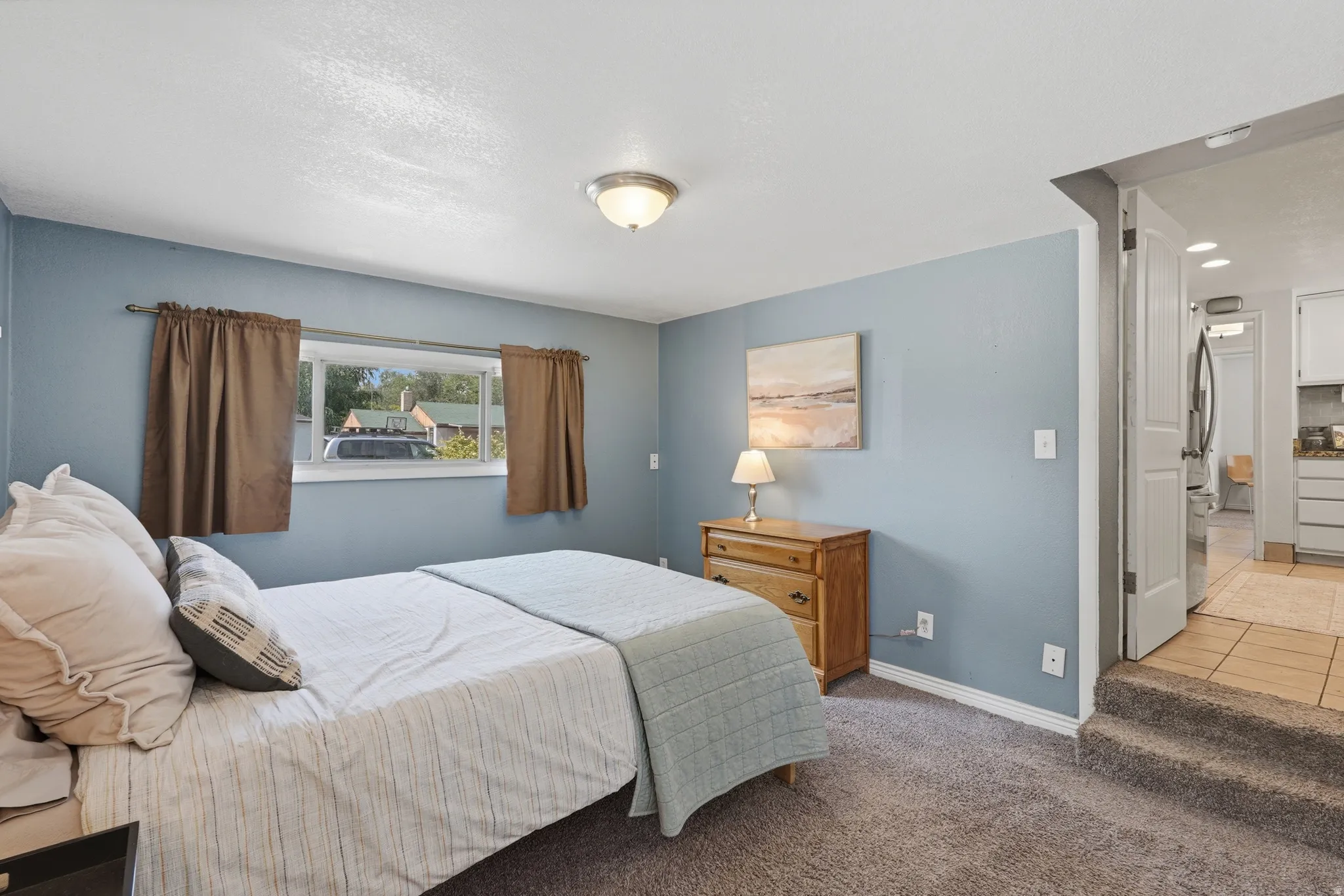 Serene primary bedroom showcasing soft blue walls, carpeted flooring, and abundant natural light from a large window with brown drapery
