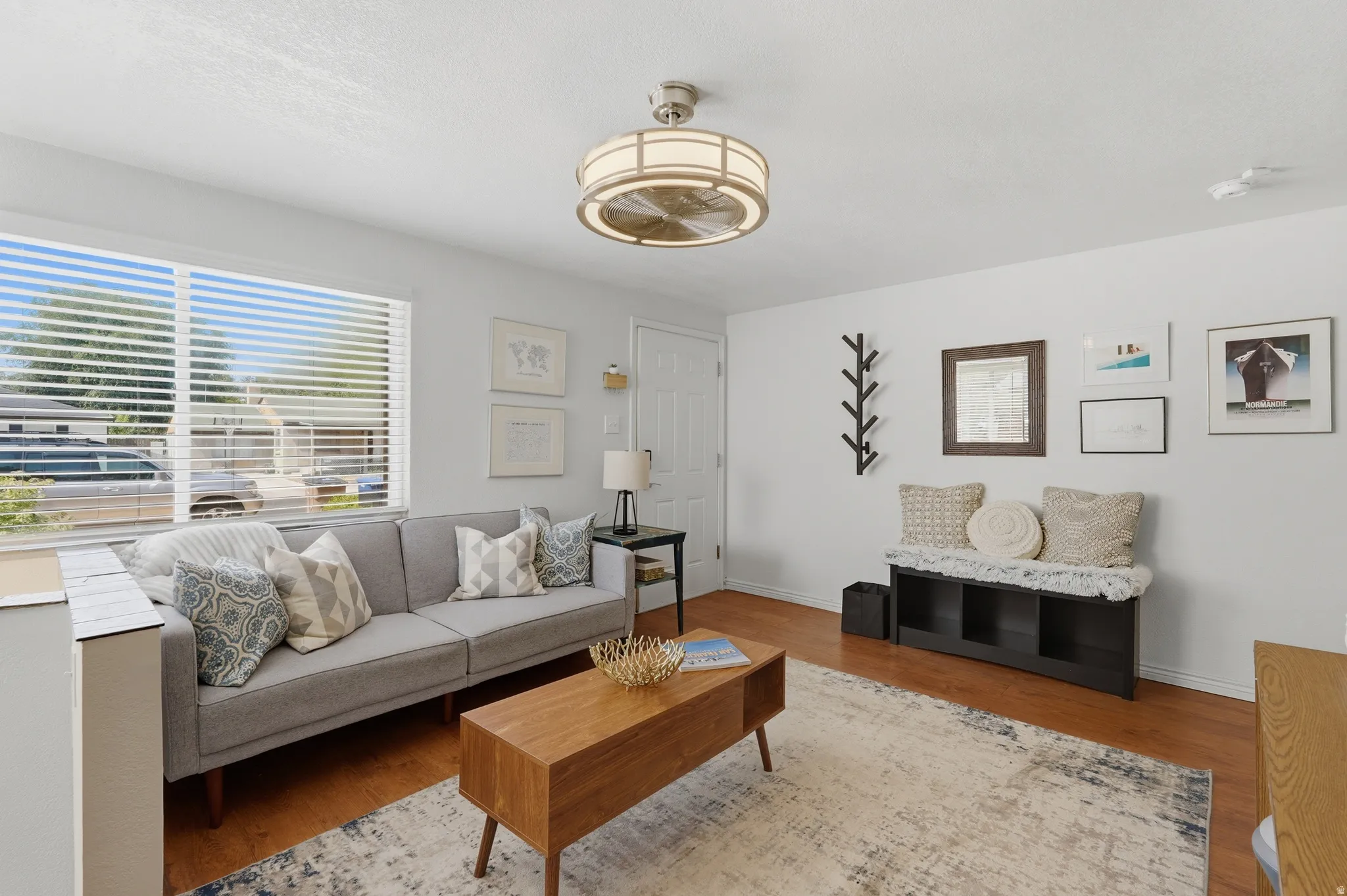 Bright living room featuring hardwood flooring, a ceiling-mounted light fixture with brushed brass details, and large windows with horizontal blinds