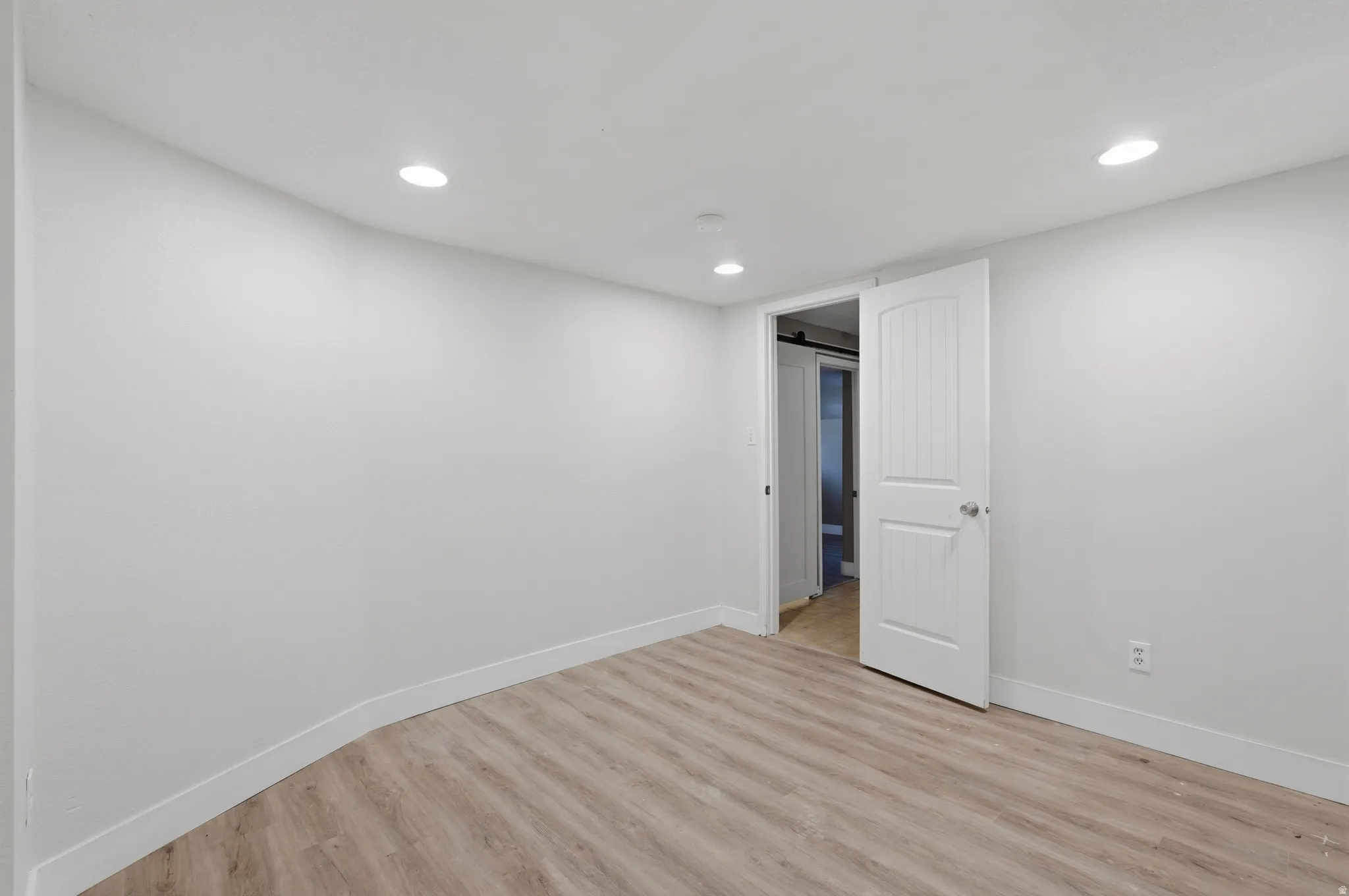 Bright, open bedroom featuring recessed ceiling lighting, pristine white walls, wood-finish flooring throughout, and white six-panel closet doors with direct access to an ensuite bathroom