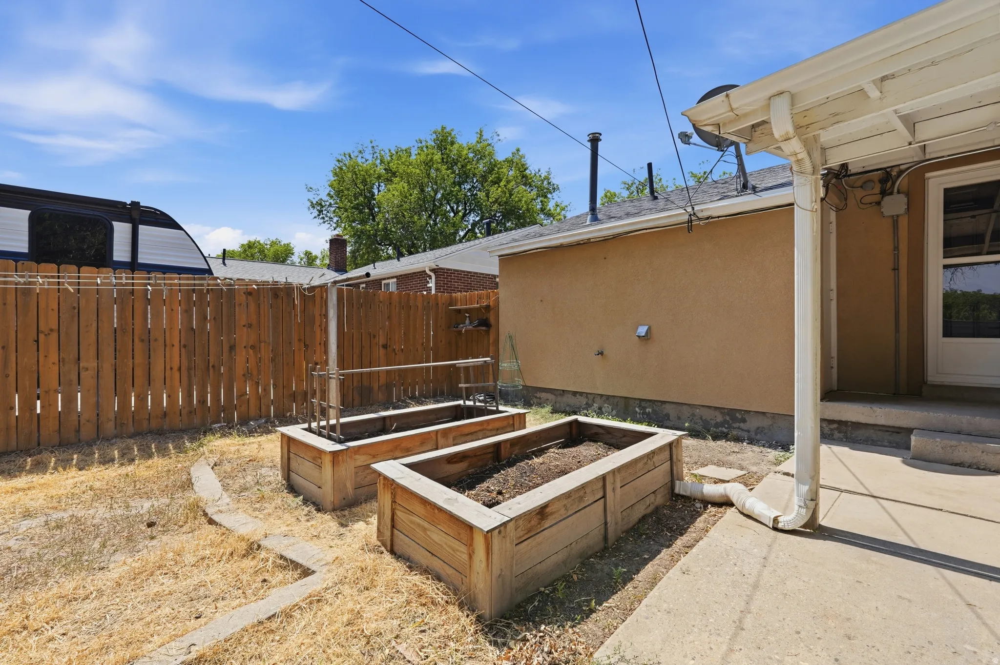 Backyard oasis featuring two spacious raised garden beds with wood-finish frames, mature shade trees, wooden privacy fencing, and a covered patio structure with pergola detailing