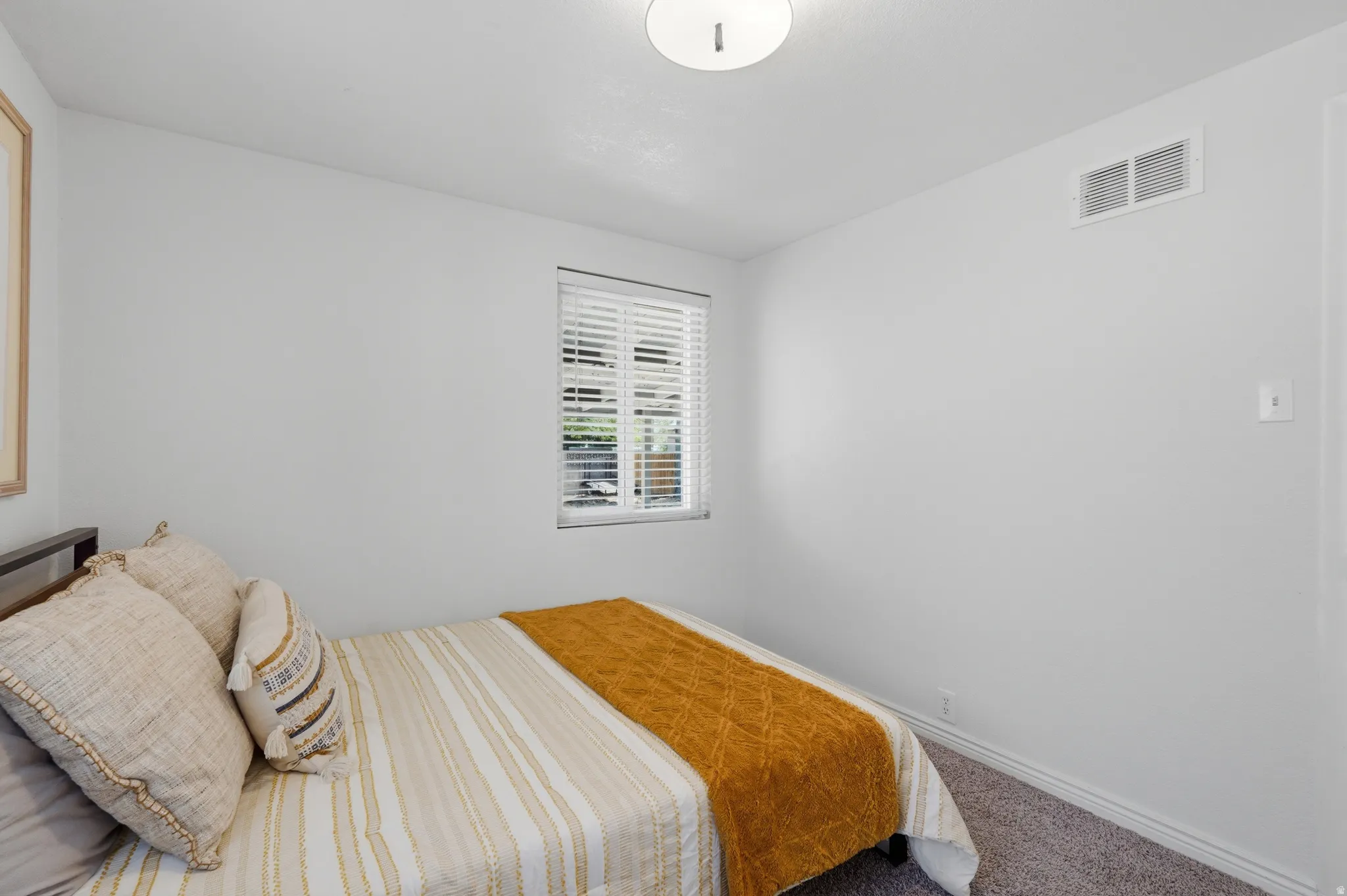 Comfortable secondary bedroom featuring a large window with plantation shutters, neutral white walls, and carpeted flooring for warmth and comfort