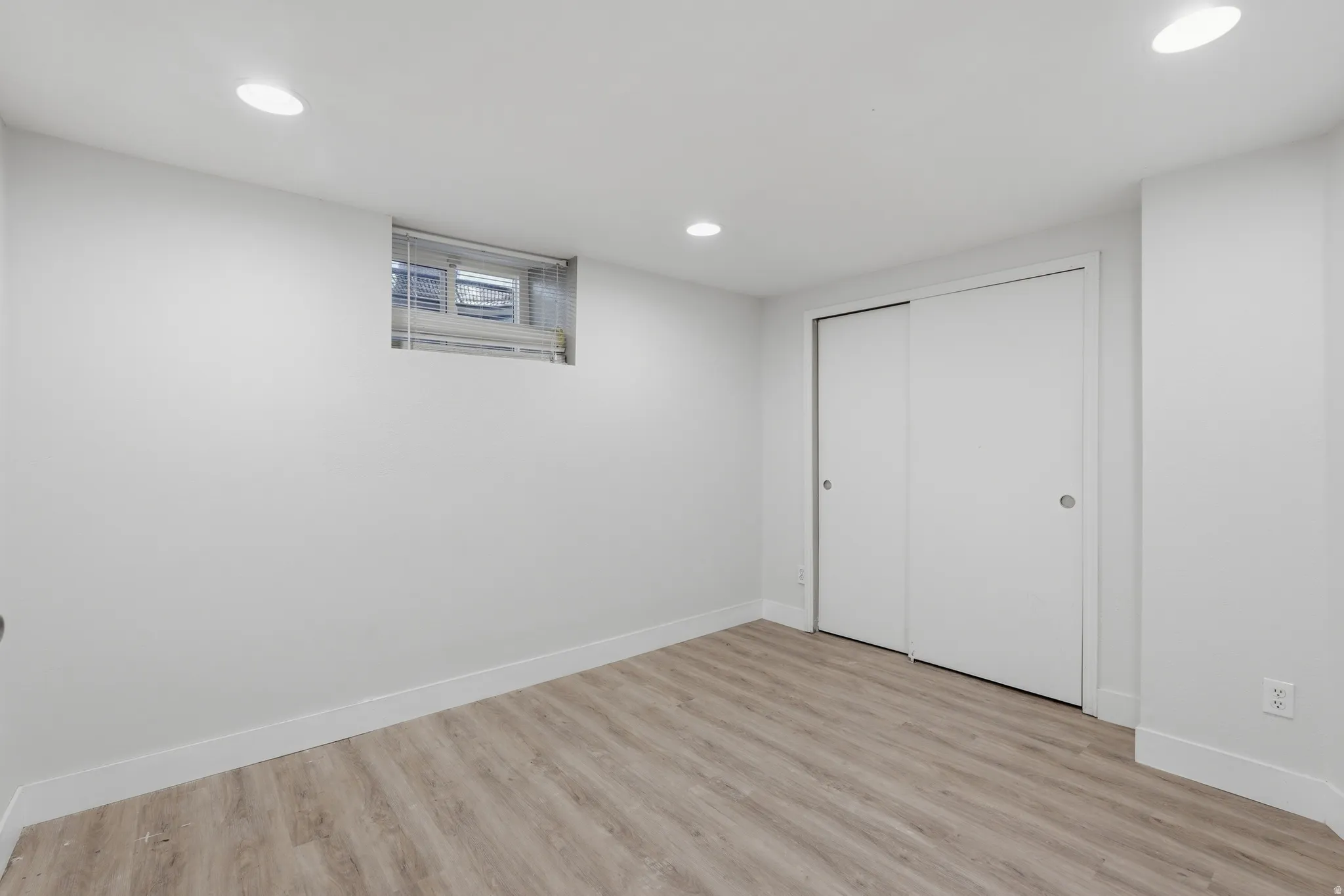 Finished basement room featuring wood-finish flooring, recessed ceiling lighting, a casement window with blinds, and dual closet doors for convenient storage
