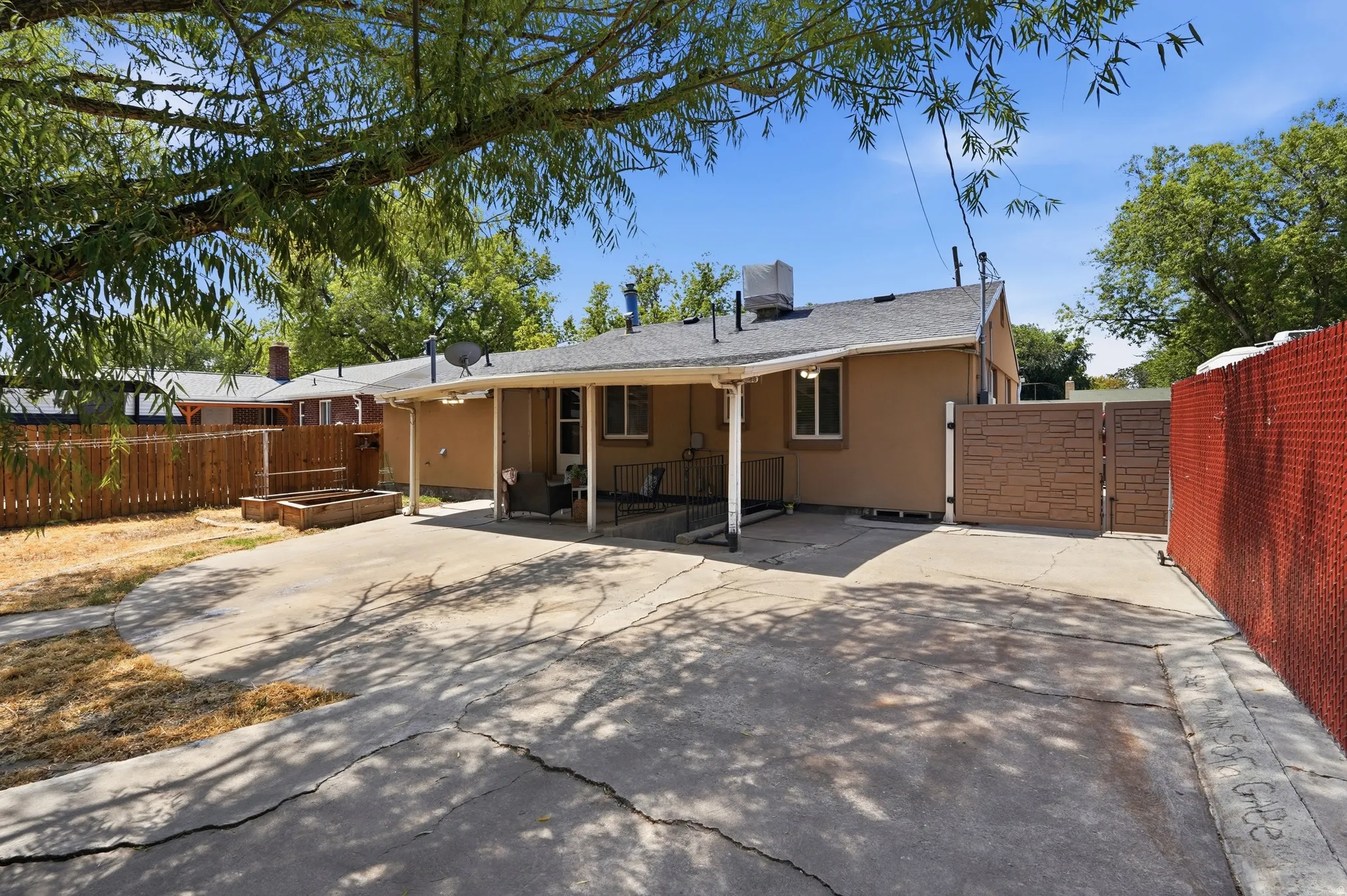 Spacious backyard featuring mature shade trees, concrete patio with attached covered structure, wooden privacy fencing, and single-story stucco residence with metal roof