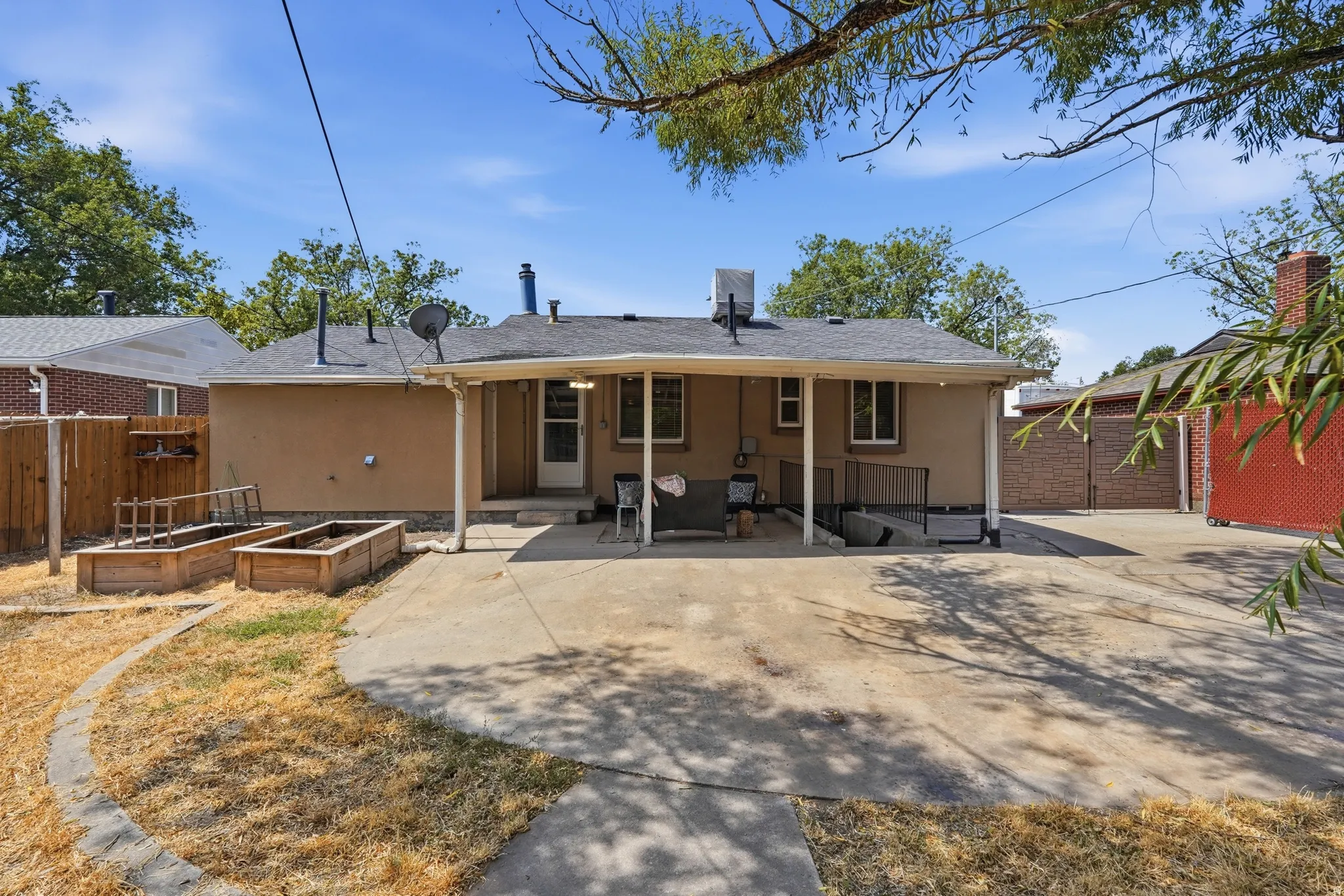 Expansive concrete patio with covered awning, raised garden beds, and a perimeter fence