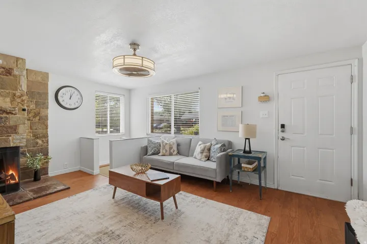 Stone-clad fireplace with hearth, wood-finish flooring, two large windows with blinds, recessed entry area, and a ceiling fan with integrated lighting