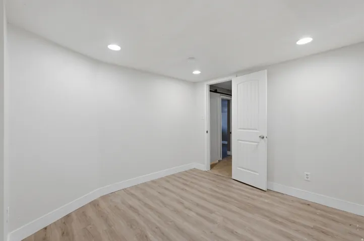 Bright, open bedroom featuring recessed ceiling lighting, pristine white walls, wood-finish flooring throughout, and white six-panel closet doors with direct access to an ensuite bathroom