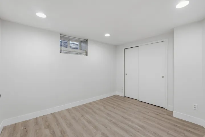 Finished basement room featuring wood-finish flooring, recessed ceiling lighting, a casement window with blinds, and dual closet doors for convenient storage