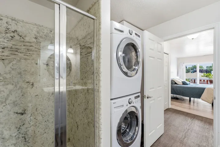Glass-enclosed shower with a tiled surround, stacked laundry appliances, and wood-finish flooring