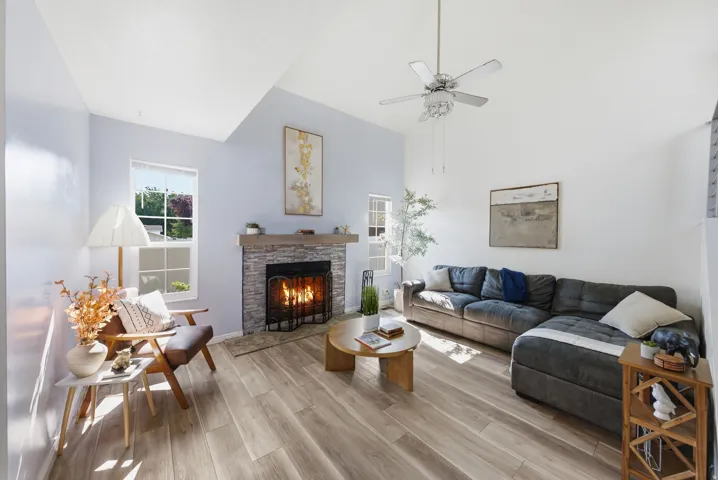 Vaulted ceilings, wood-finish flooring, stone-clad fireplace with wood mantel, a ceiling fan, and multiple windows