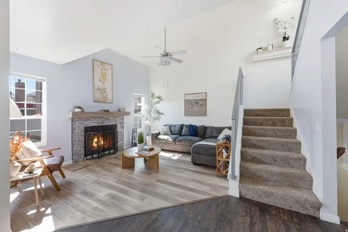 Vaulted ceiling with ceiling fan, stone-clad fireplace with mantel, wood-finish flooring, and carpeted staircase with metal railings