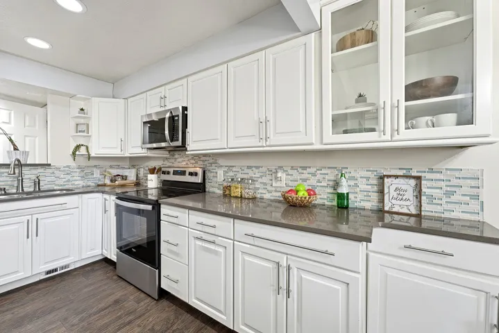 Bright, modern kitchen featuring white shaker-style cabinetry with glass-front upper cabinets, stainless steel appliances including a slide-in range and microwave, gray quartz countertops, and a geometric tile backsplash