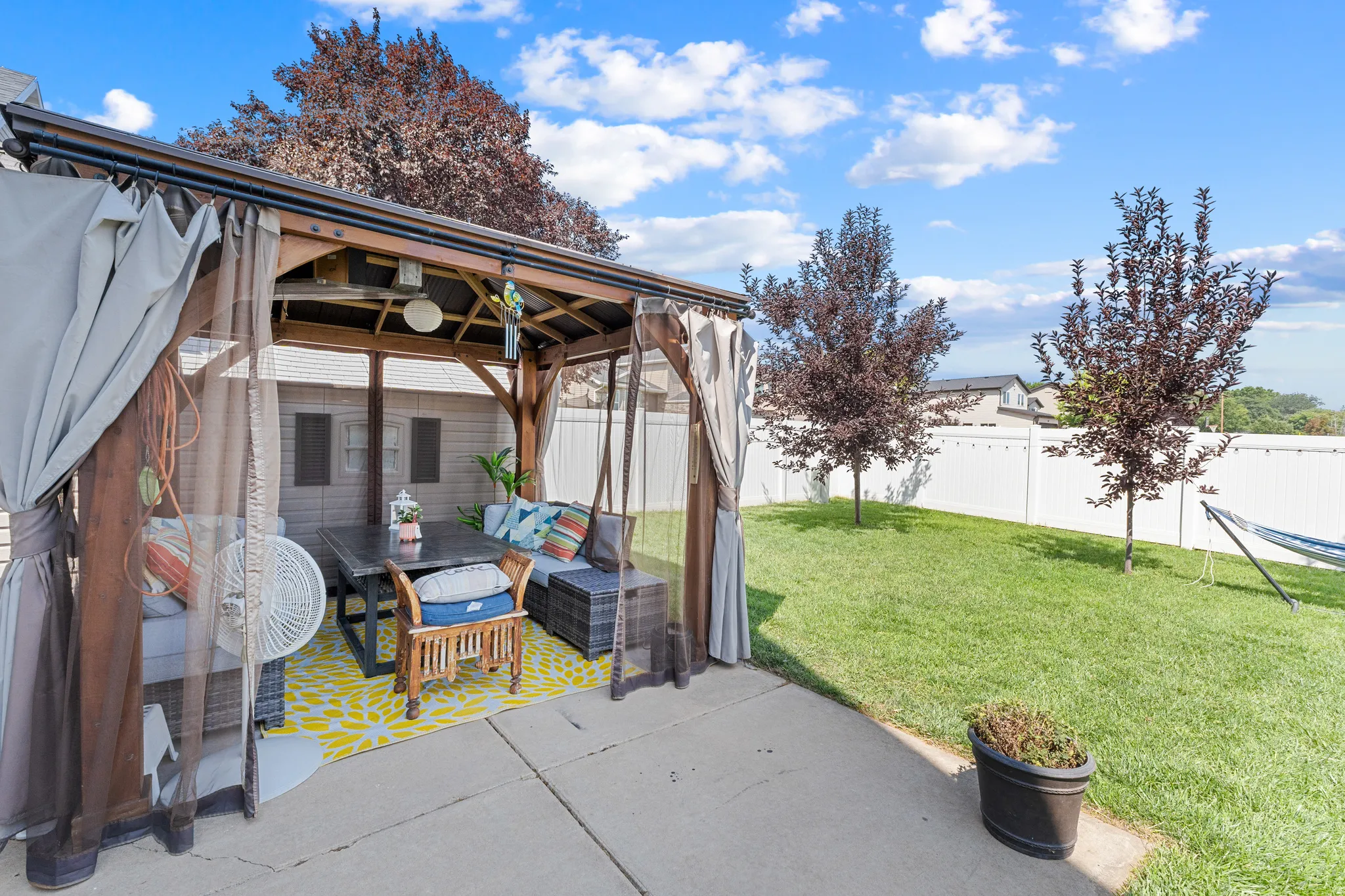 Outdoor gazebo with privacy screens, concrete patio, expansive lawn, white privacy fence, and mature trees