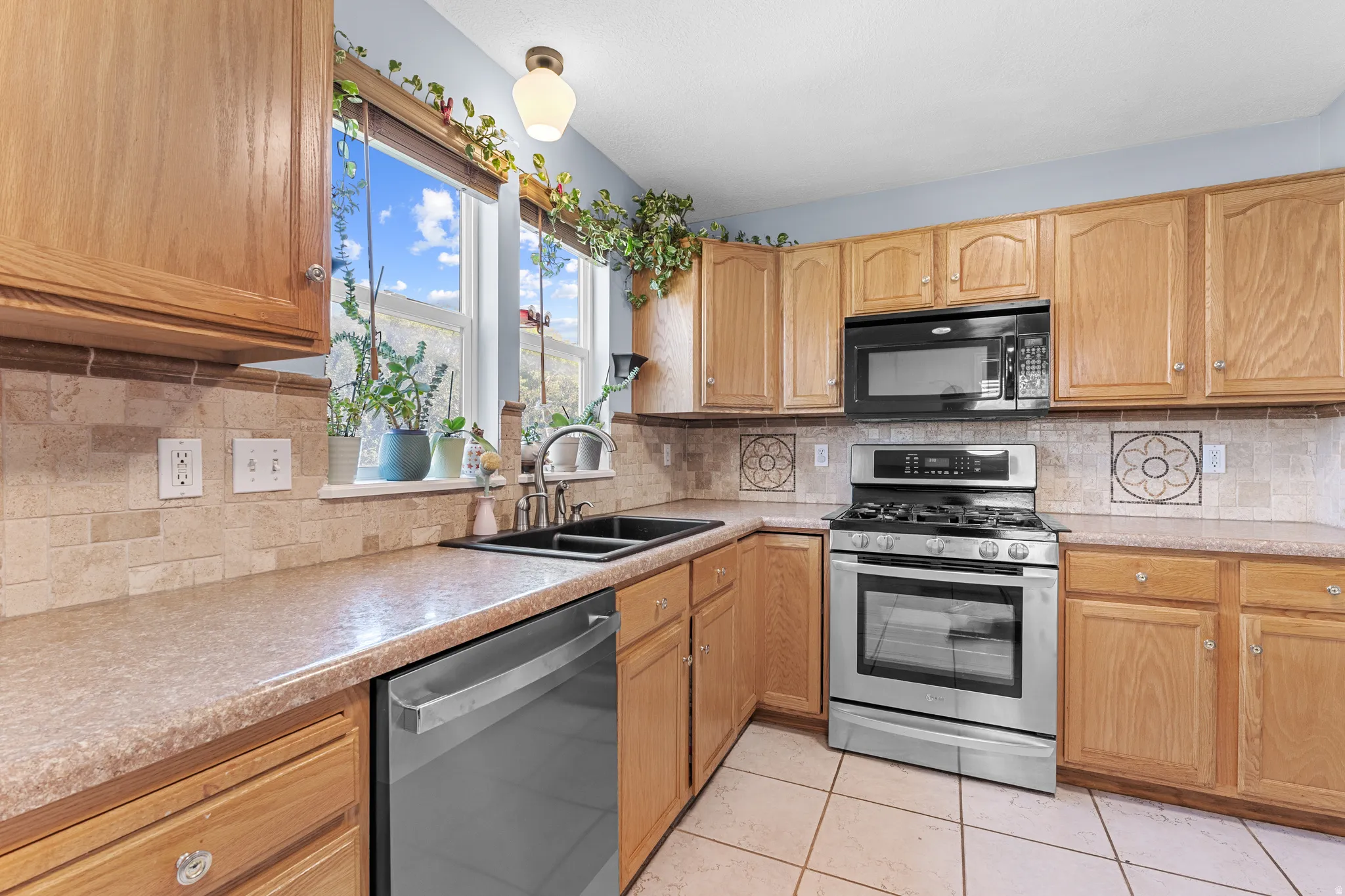 Kitchen featuring light wood cabinetry, a tile backsplash with decorative accents, a stainless steel gas range, and a dual basin sink