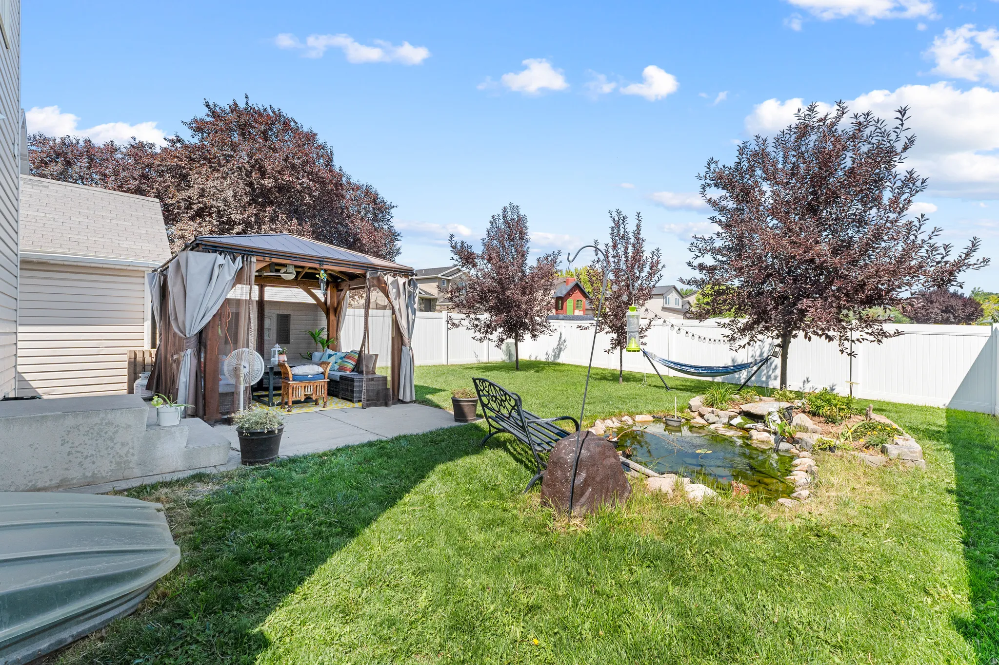 Expansive lawn with a white privacy fence, mature trees, a built-in pond feature, and a patio with a gazebo