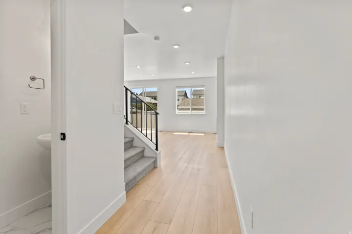 Recessed lighting, light wood-finish flooring, carpeted staircase with black metal railing, white walls, and a half-bath with a pedestal sink and marble-look tile flooring