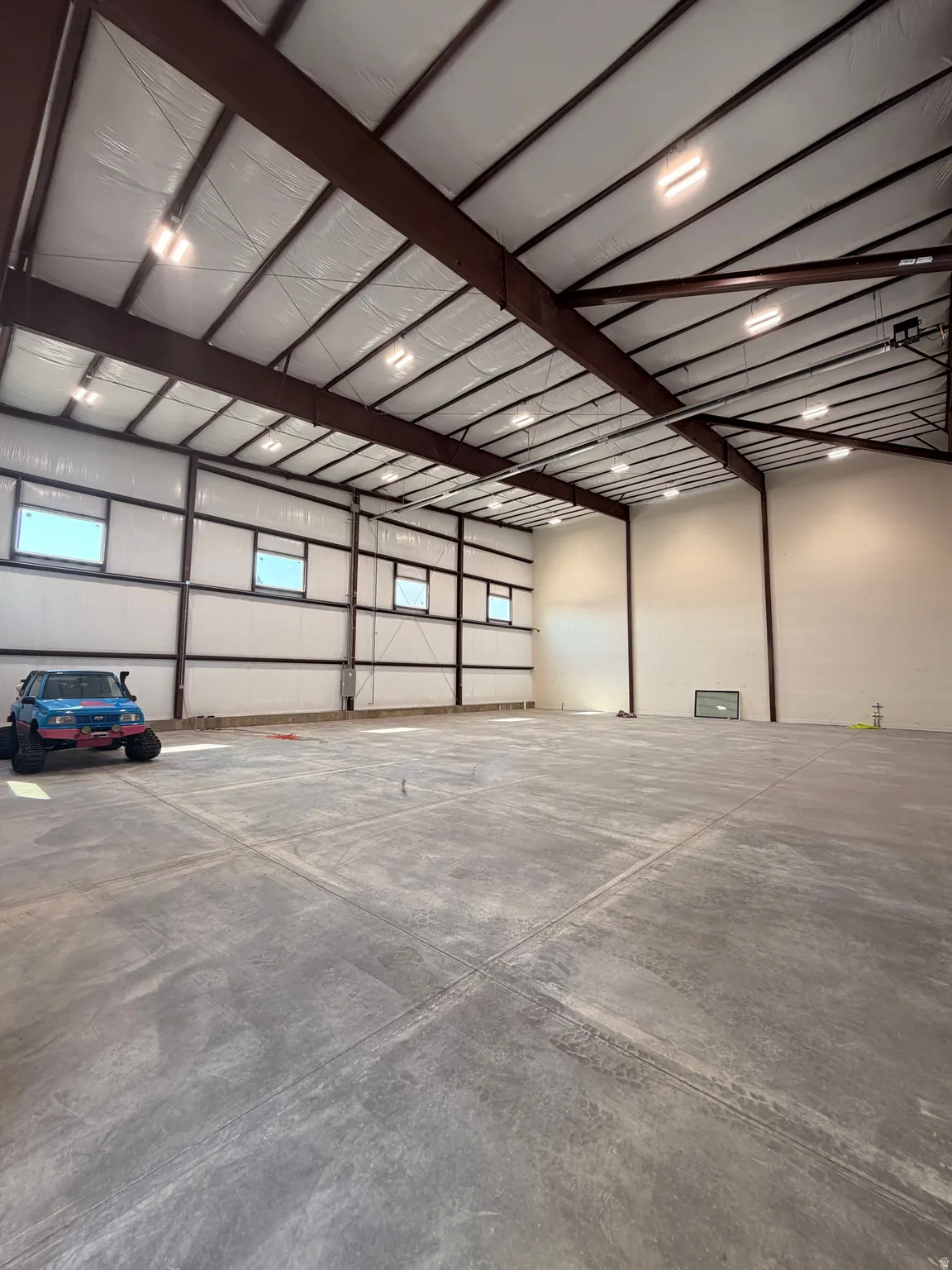 Spacious warehouse featuring a concrete floor, high ceilings with exposed steel beams, multiple windows providing natural light, and overhead lighting fixtures