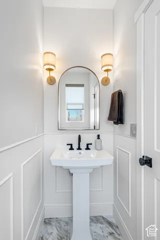 Elegant powder room featuring a pedestal sink with polished chrome faucet and fixtures, arched mirror with brushed metal frame, dual brass wall sconces with frosted glass shades, white shiplap wainscoting, and marble-look tile flooring