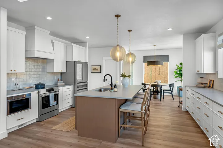 Kitchen featuring white shaker cabinetry, stainless steel appliances, a center island with bar seating, wood-finish flooring, and pendant lighting