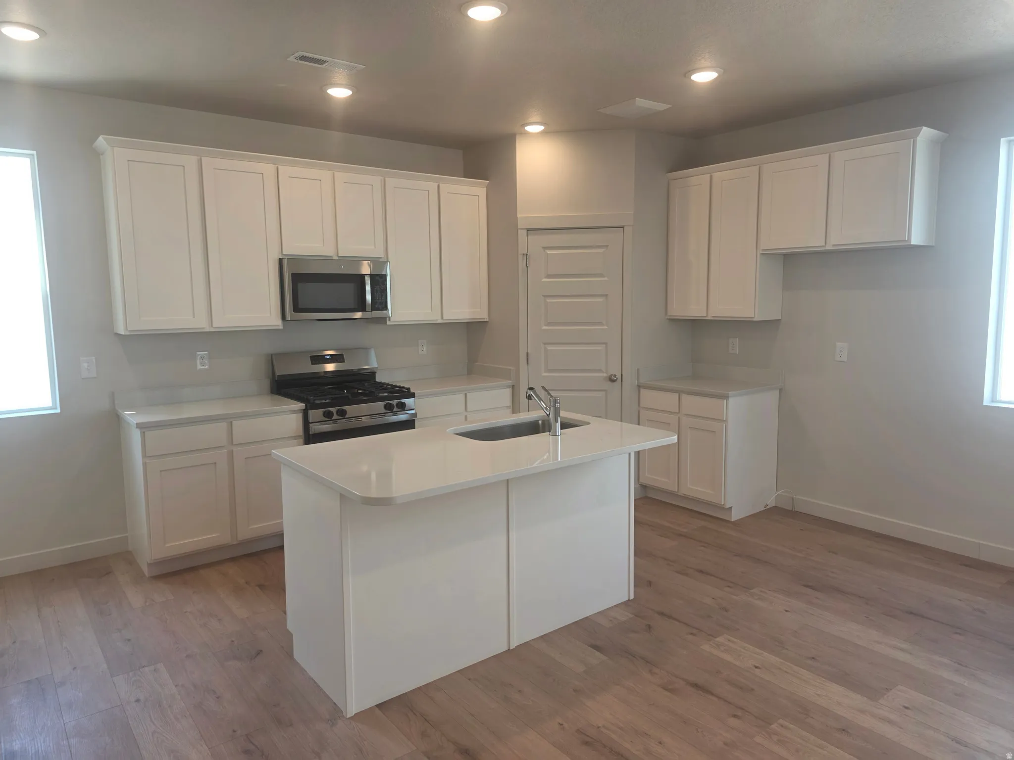 Kitchen featuring white shaker cabinetry, stainless steel appliances, a center island with sink, light countertops, and wood-finish flooring