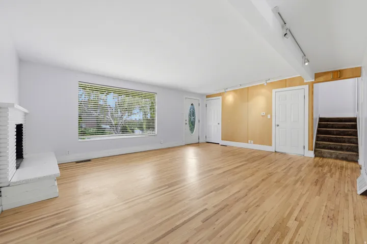 Hardwood flooring throughout, a white painted brick fireplace, a large window with horizontal blinds, a front entry door with an oval glass insert, and track lighting