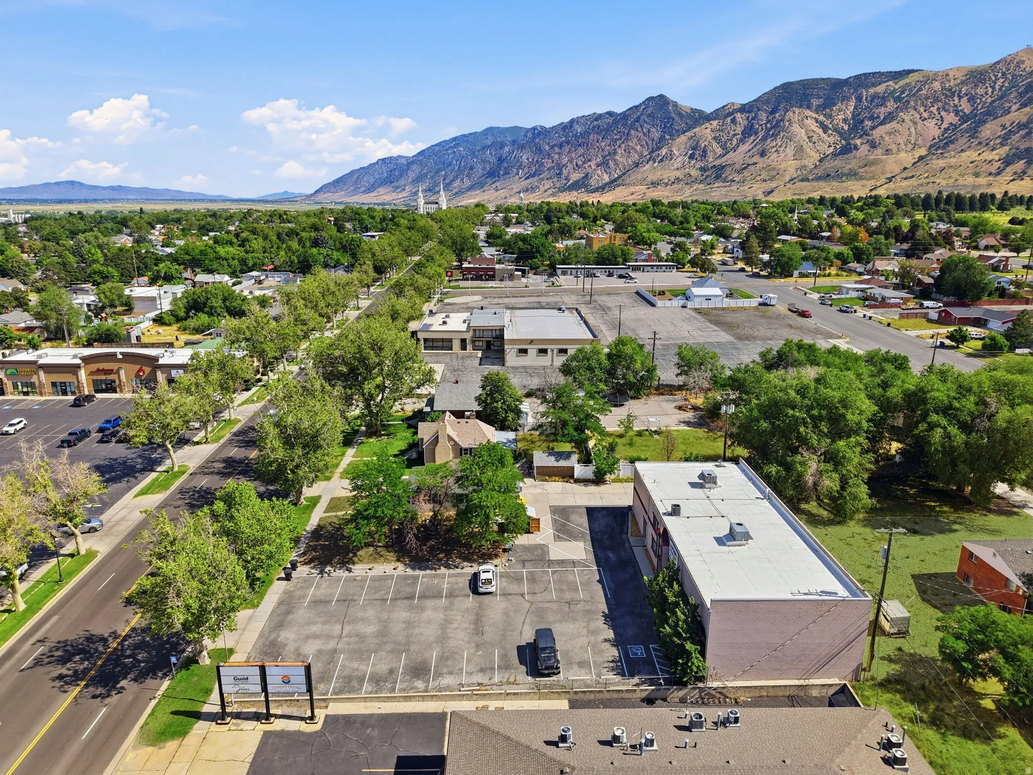 Aerial view showcasing a commercial/industrial property with a flat roof structure, expansive paved parking area, and mature landscaping throughout the grounds