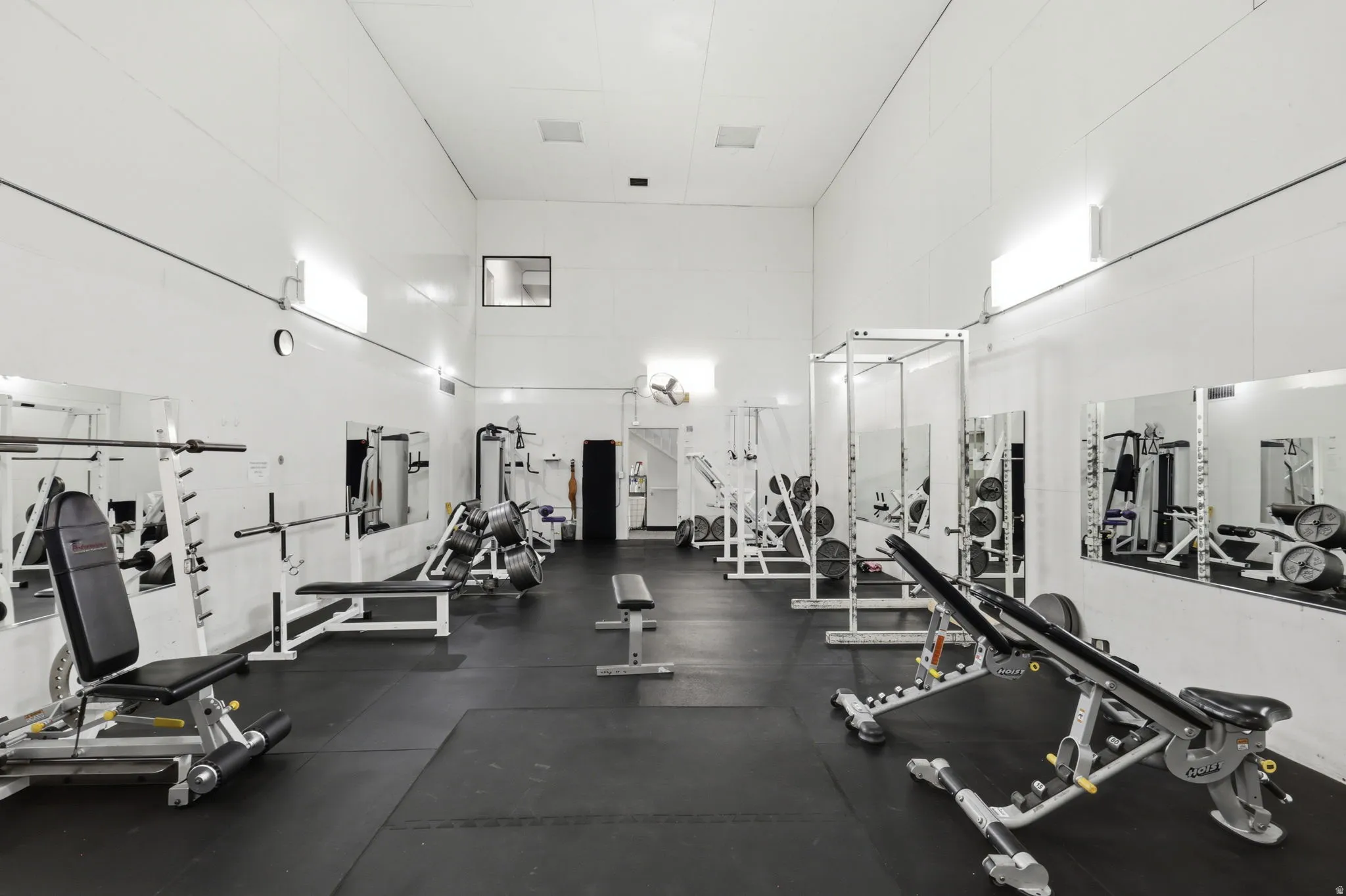Spacious recreation area featuring high ceilings, dark rubber flooring, white walls, and built-in wall mirrors