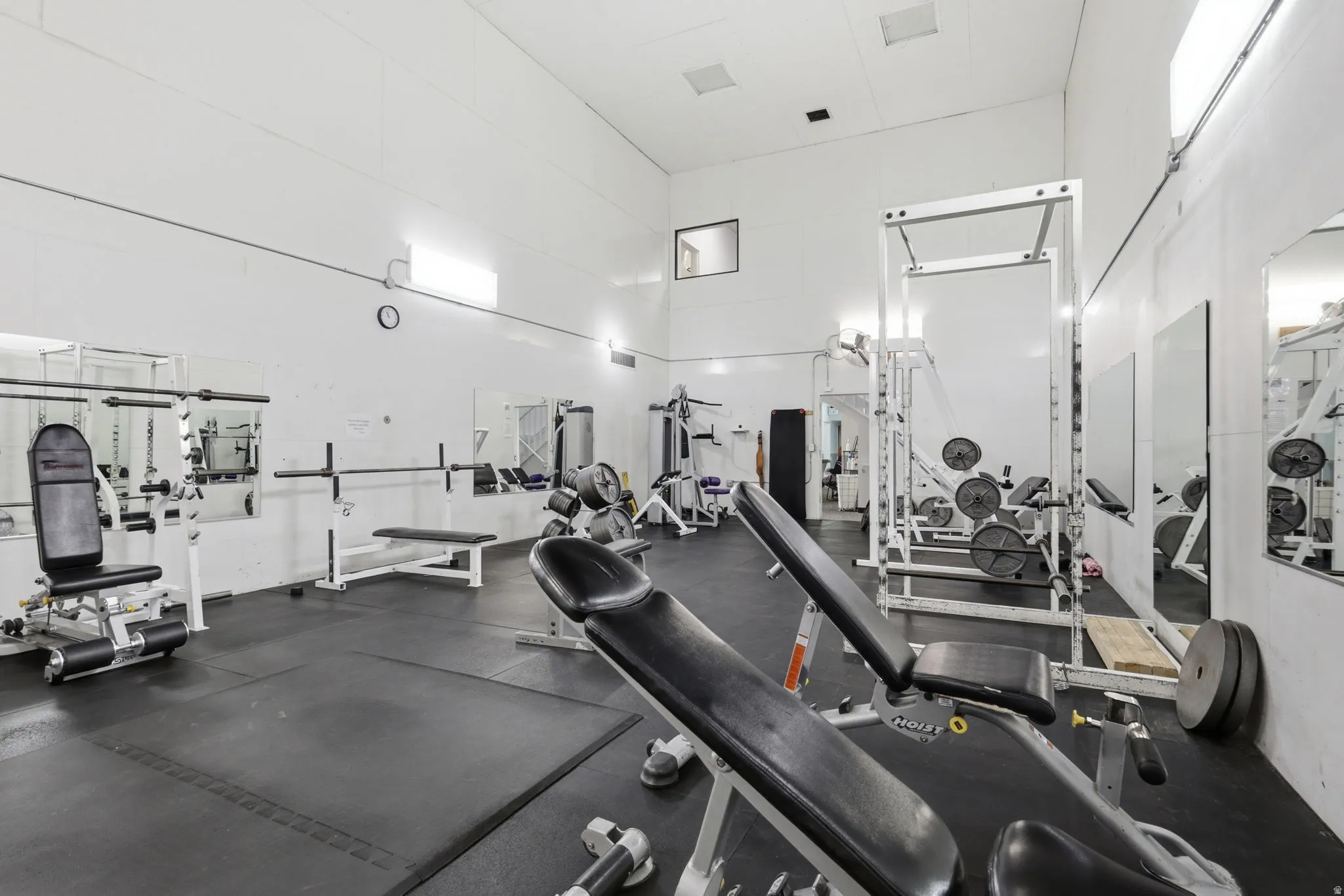 State-of-the-art fitness facility featuring multiple strength training stations, cardio equipment, mirrored walls for form assessment, rubber flooring throughout, and abundant natural lighting from skylights and recessed ceiling fixtures