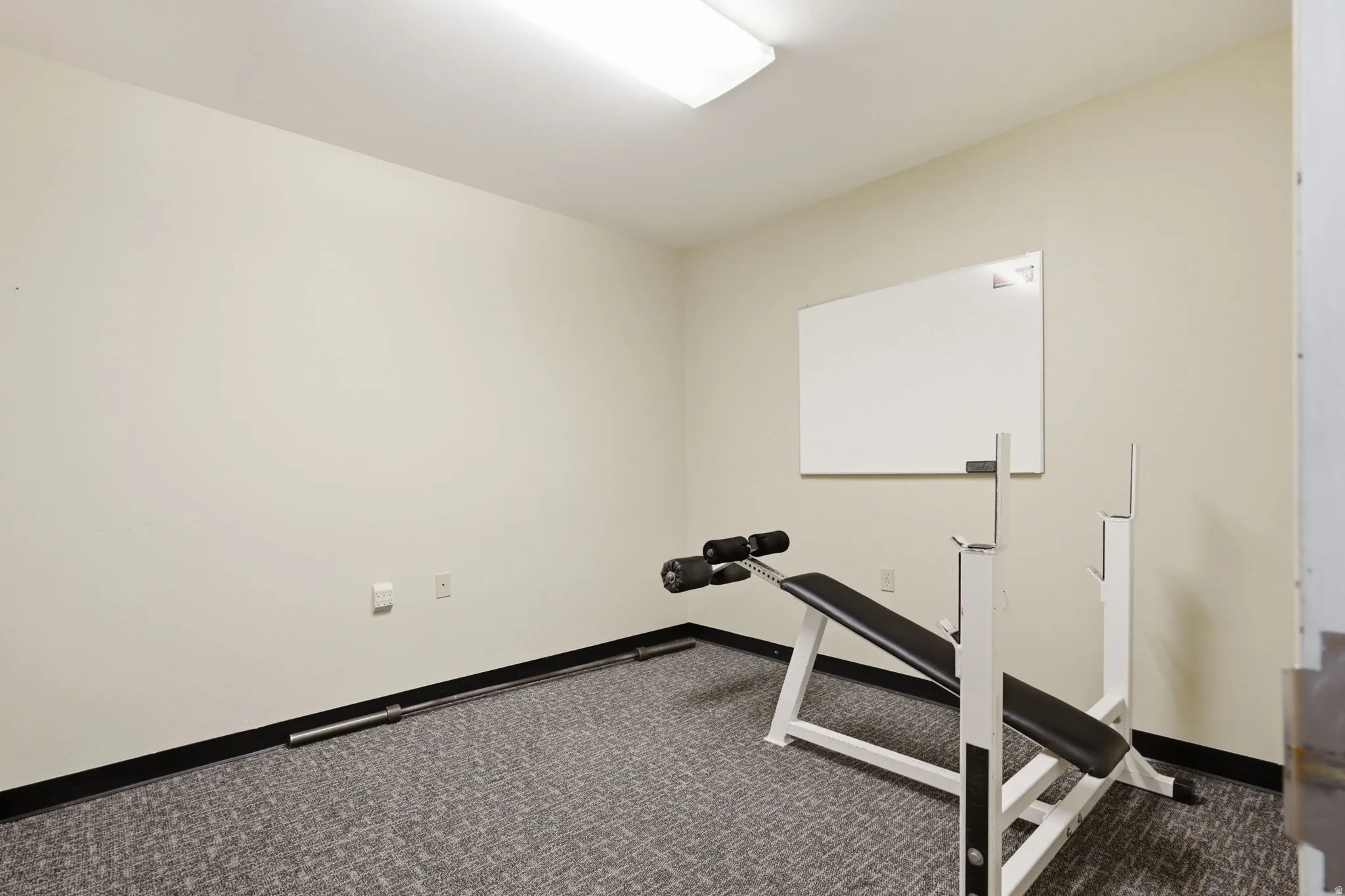 Dedicated fitness room featuring neutral wall finishes, recessed ceiling lighting, carpeted flooring with dark border trim, a weight bench with adjustable frame, and a whiteboard for workout tracking