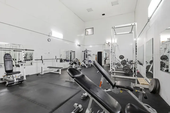 State-of-the-art fitness facility featuring multiple strength training stations, cardio equipment, mirrored walls for form assessment, rubber flooring throughout, and abundant natural lighting from skylights and recessed ceiling fixtures