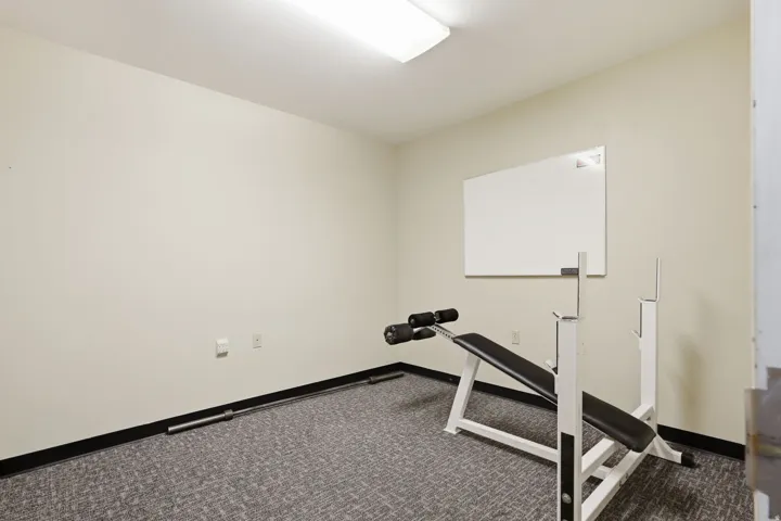 Dedicated fitness room featuring neutral wall finishes, recessed ceiling lighting, carpeted flooring with dark border trim, a weight bench with adjustable frame, and a whiteboard for workout tracking