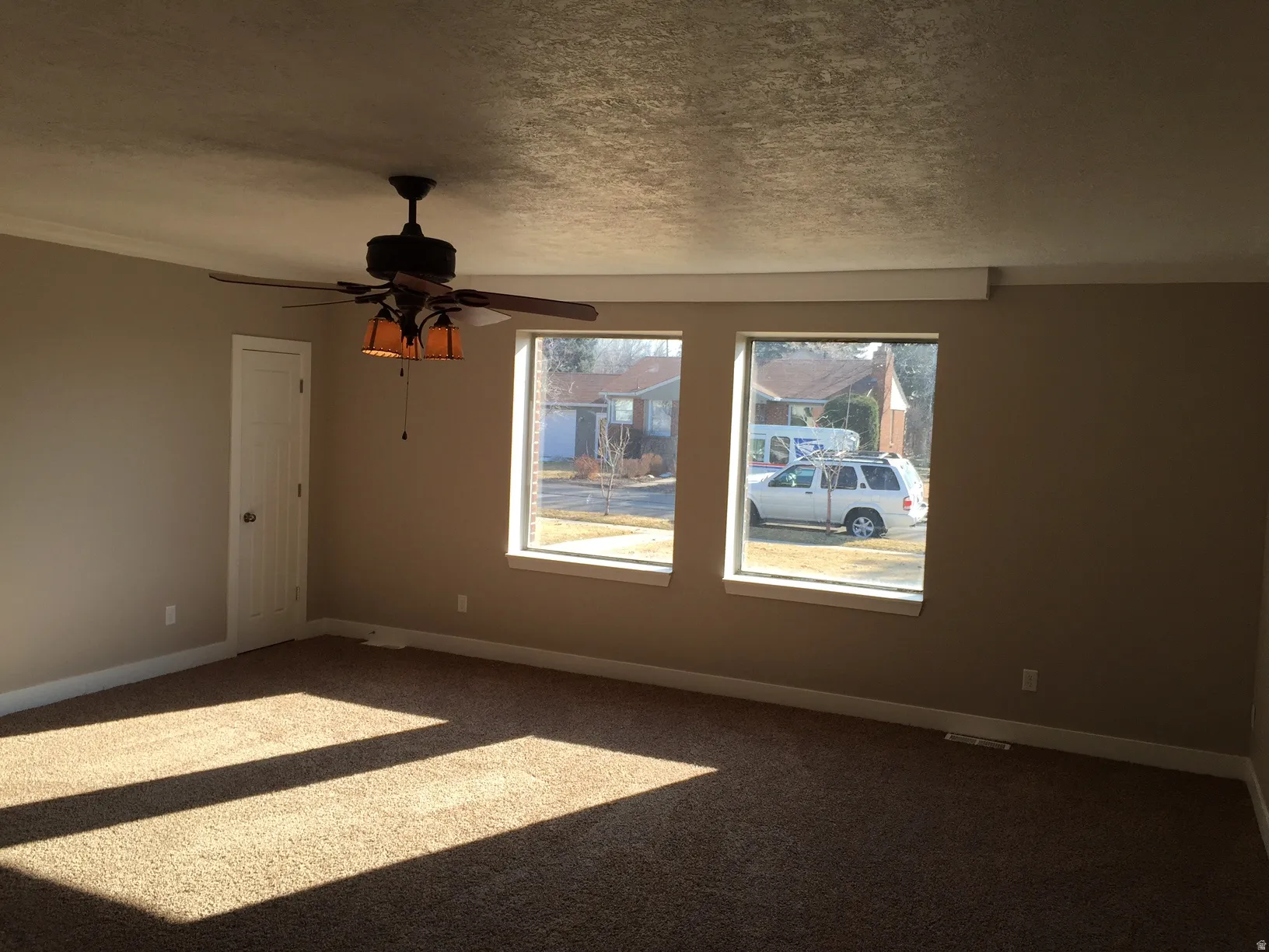 Neutral tone wall paint, textured ceiling, two large windows, ceiling fan with integrated lighting, and carpet flooring