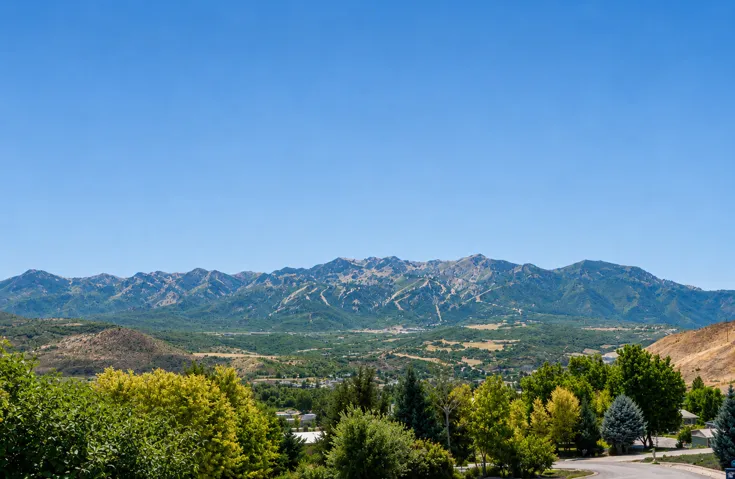 Expansive mountain range backdrop with distant ski slopes, lush green foliage, and residential areas in the valley