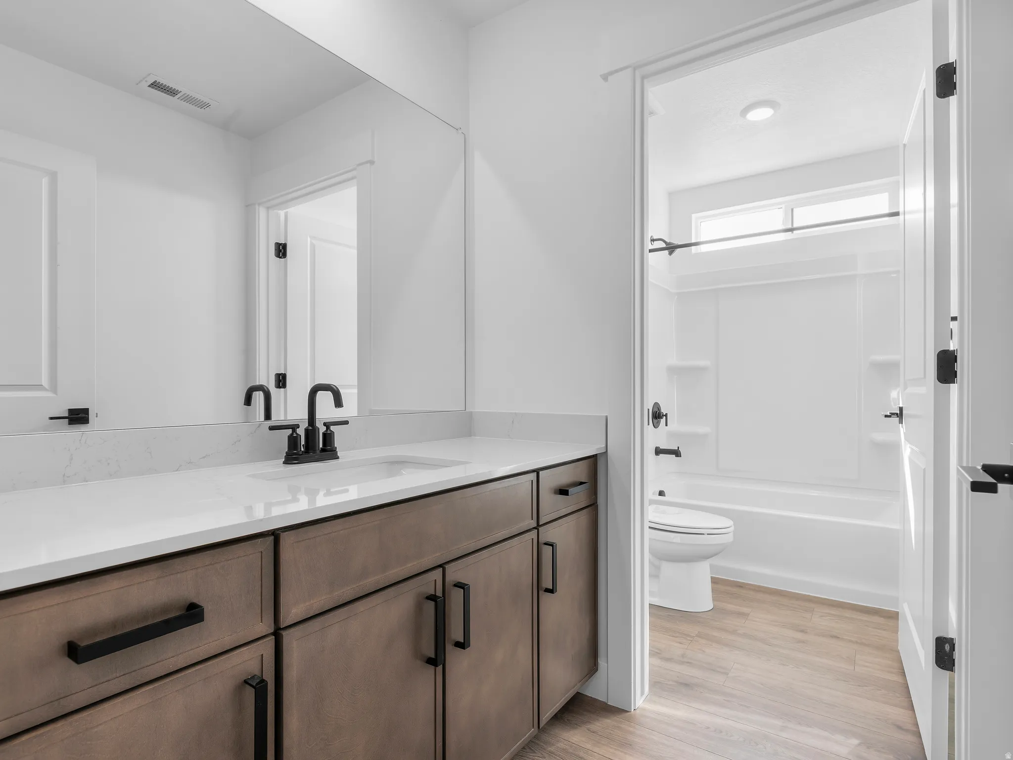 White countertop with integrated sink, black hardware, wood-finish cabinetry, full-width mirror, and a full bath with a window