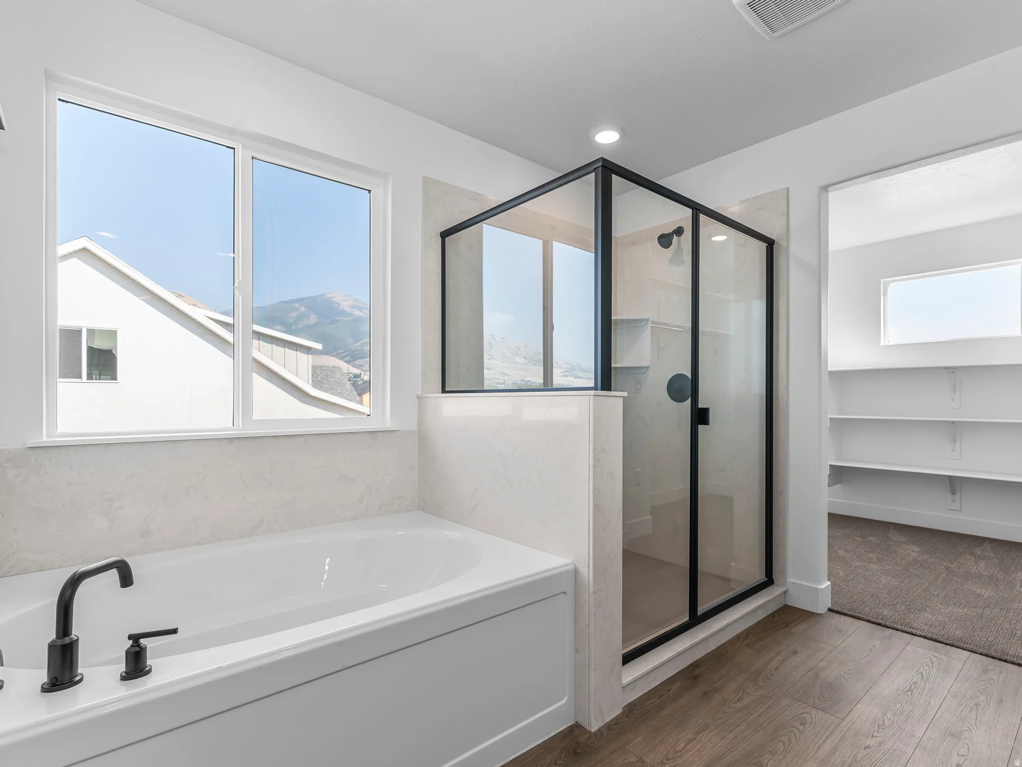 Soaking tub with matte black fixtures, an enclosed glass shower with black framing, wood-finish flooring, and a large window